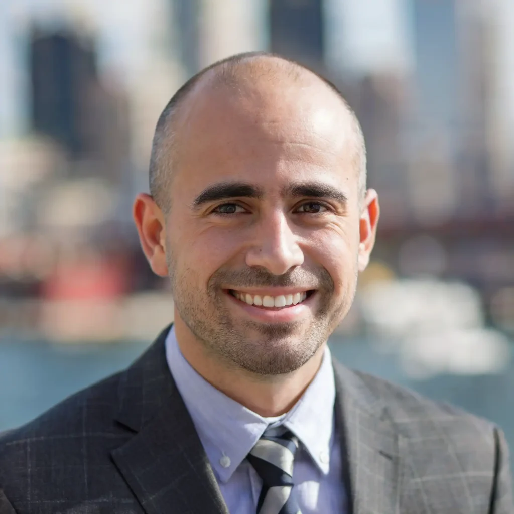 A man in a suit and tie smiles outdoors with a blurred cityscape in the background.